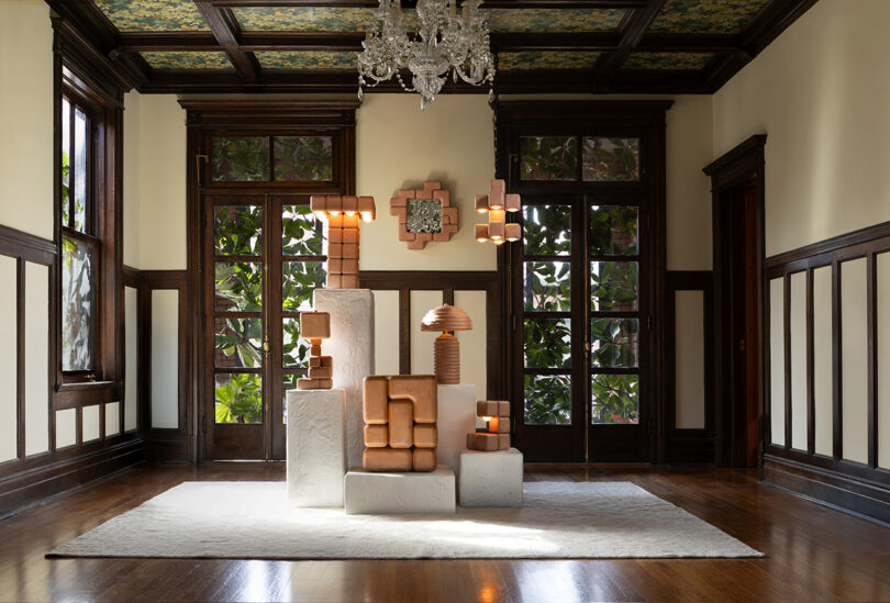A grouping of geometric sculptural lamps and decor is arranged on white blocks in the center of a formal wood-paneled room with large windows, a chandelier, and a white rug.