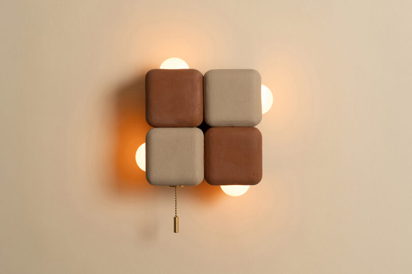 A wall-mounted light fixture with four rounded square panels in brown and beige, four exposed bulbs at the corners, and a pull chain hanging below.