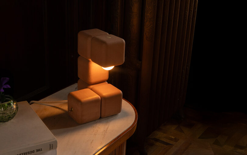 A table lamp with a design of stacked, cube-shaped blocks emits warm light on a marble-topped table next to a book and a small glass vase with a purple flower.