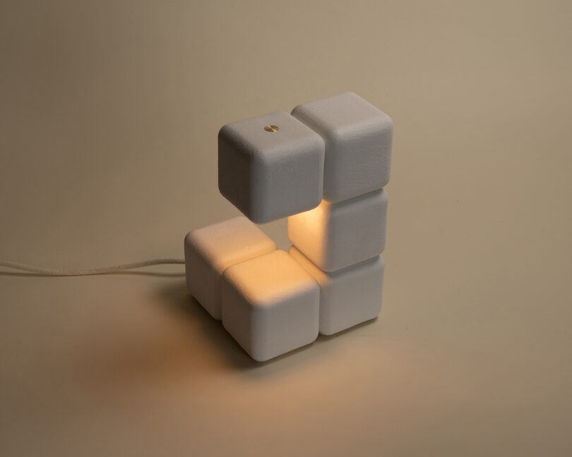 A modern lamp made of stacked white cubes emits a warm light from its center, placed on a beige background with a visible power cord.