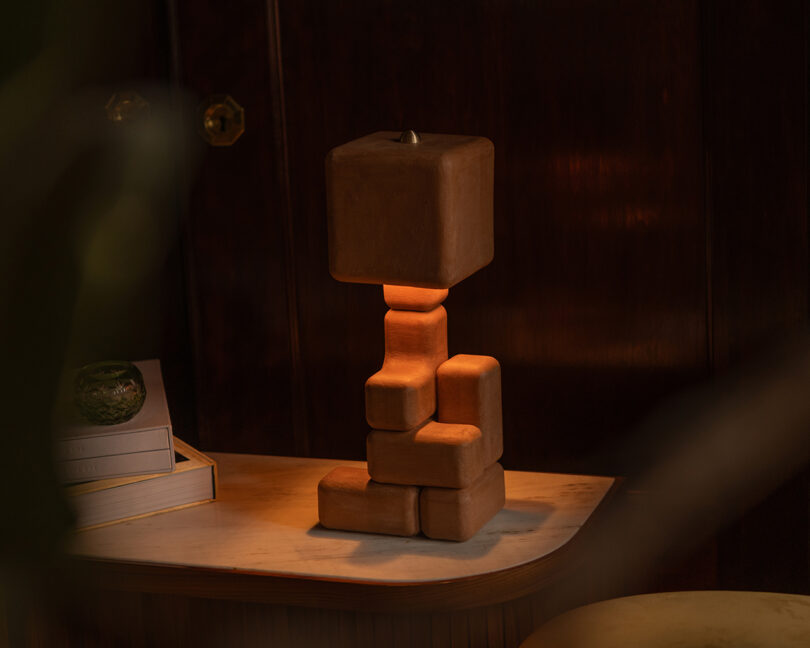 A sculptural table lamp with a stacked, block-like wooden base and a matching square wooden shade sits on a marble-top table beside books and a small decorative object.
