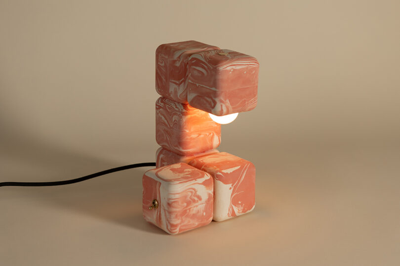 A table lamp made of stacked, marbled pink and white ceramic cubes with a visible light bulb and a black power cord.