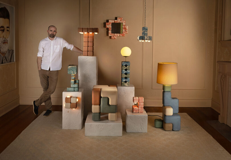 A man stands beside geometric, modern lamps and sculptures displayed on textured white pedestals in a warmly lit room with beige walls and carpet.