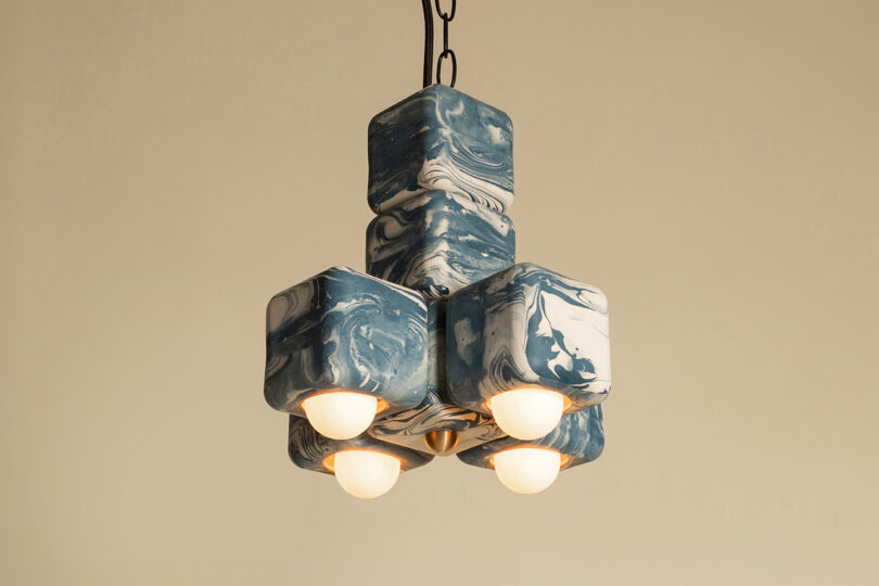 A modern pendant light fixture with six cube-shaped, blue and white marbled casings, each housing a round bulb, hangs from a black chain against a beige background.