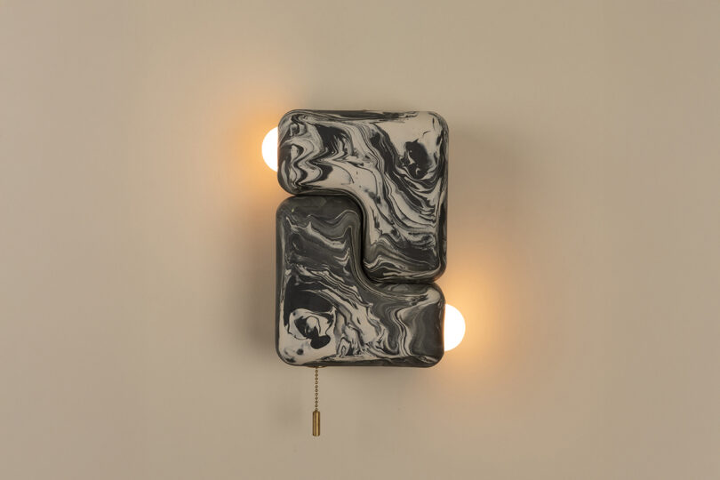 Wall-mounted light fixture with two glowing bulbs and marbled black-and-white rectangular covers, featuring a pull chain hanging below.