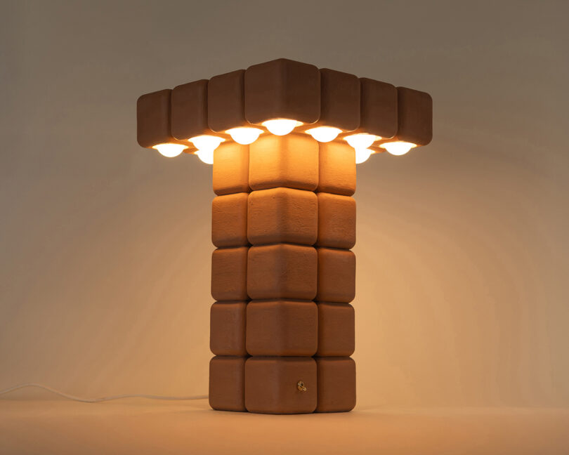 A lamp with a column and top made of stacked, rounded brown blocks, emitting warm light from bulbs arranged around the edges of the top.
