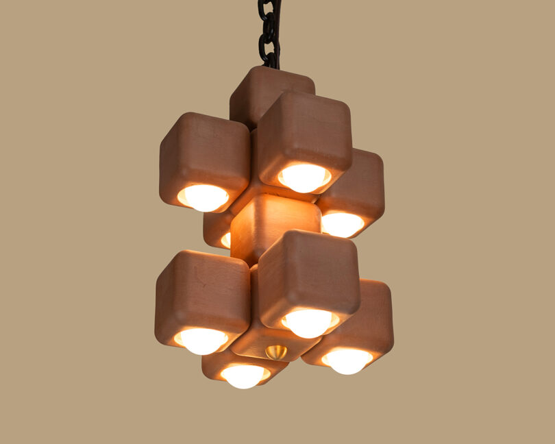 A geometric pendant light fixture with nine cube-shaped modules, each containing a lit bulb, suspended by a black chain against a beige background.