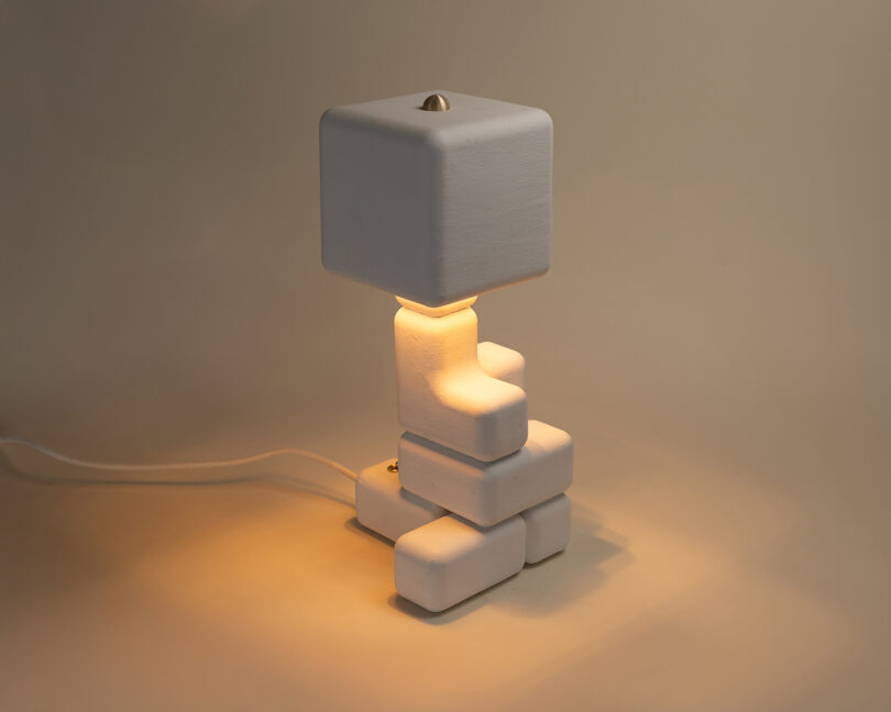 A modern table lamp with a square shade and stacked geometric white base, emitting a warm light, set against a plain background.
