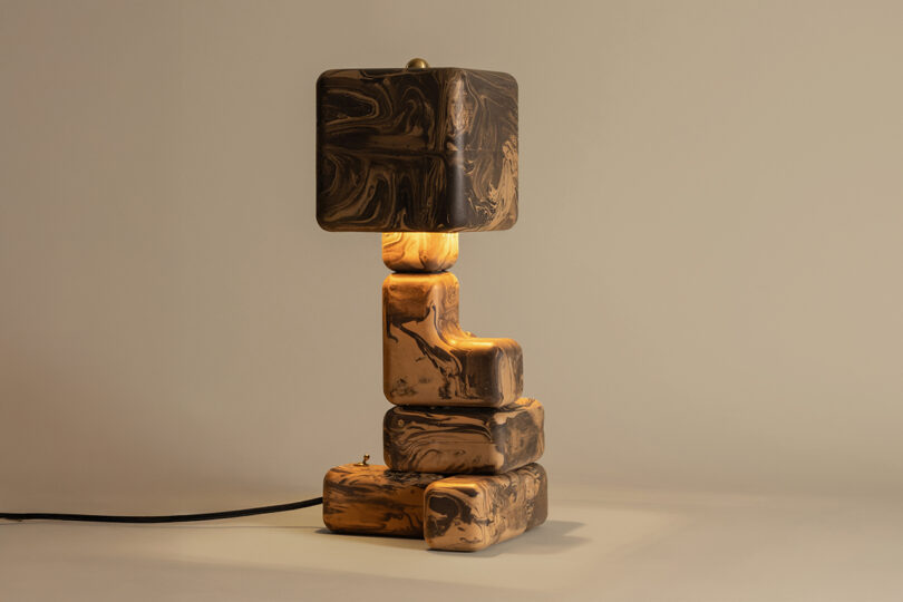 A sculptural table lamp with a black and brown marbled pattern, featuring geometric stacked shapes and a matching square lampshade, sits on a plain surface.
