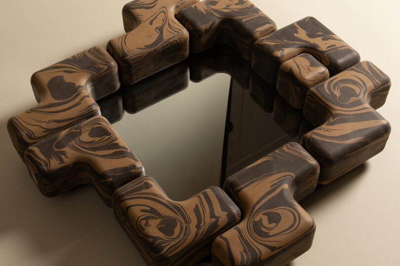 A square mirror with an irregular, chunky frame featuring a brown and black woodgrain pattern sits on a beige surface.