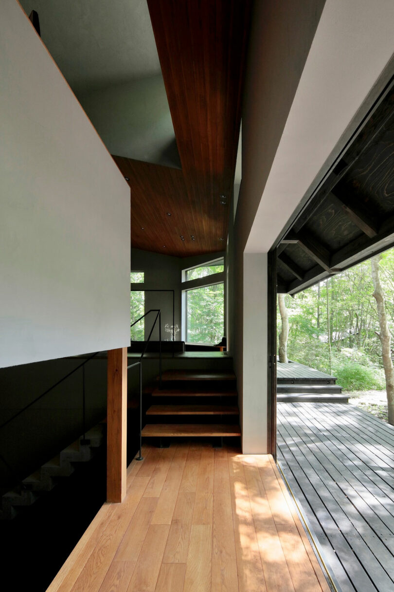 Interior view of a modern home with wooden floors and ceilings, a staircase, large windows, and an open sliding door leading to a dark wood deck surrounded by trees.