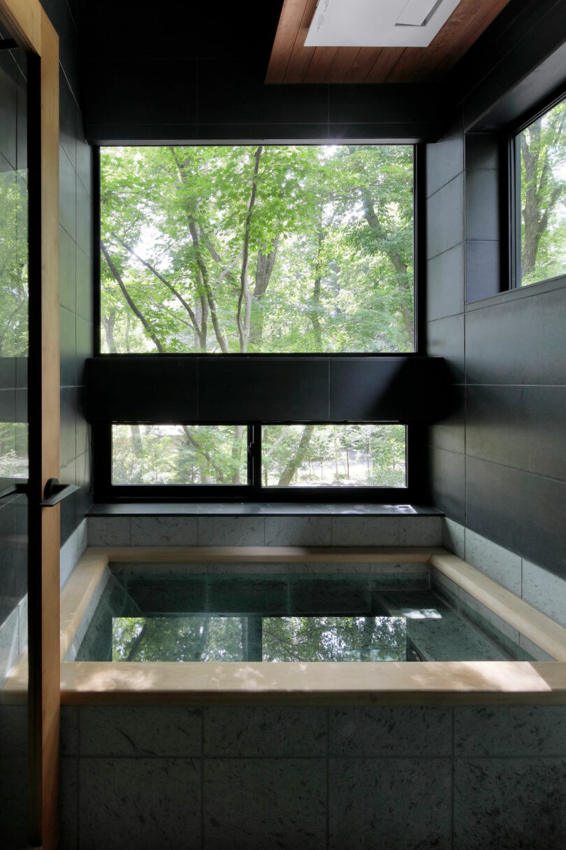 A modern indoor soaking tub with clear water is positioned by large windows overlooking green trees and natural outdoor scenery.