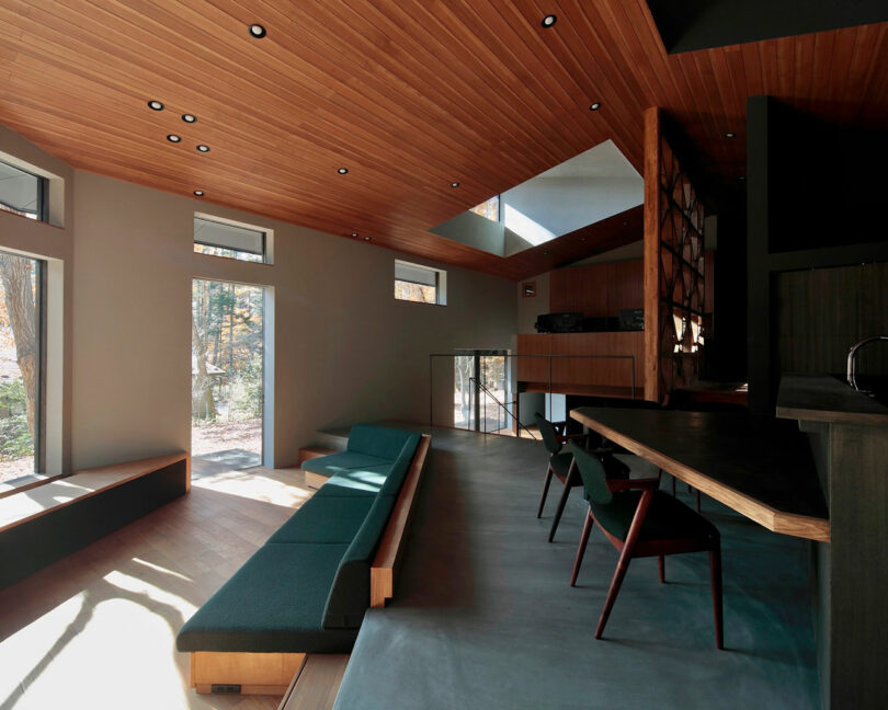 Modern living room with wood-paneled ceiling, large windows, minimalist built-in seating, dark green chairs, and an open layout with natural light.
