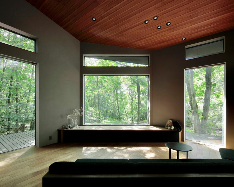 Modern living room with large windows showing a forest view, wooden ceiling, light wood floor, and minimalist furniture including a bench, sofa, and round table.