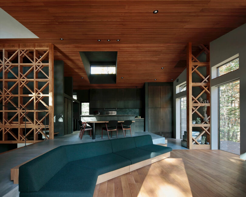 Modern open-plan living space with a dark green sectional sofa, wood ceiling and floors, geometric wood partitions, and a minimalist kitchen in the background. Natural light fills the room.