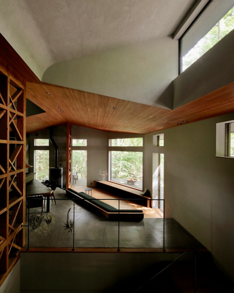 Spacious modern living room with large windows, wooden ceiling, minimalist furniture, and open shelving, overlooking a green outdoor view.