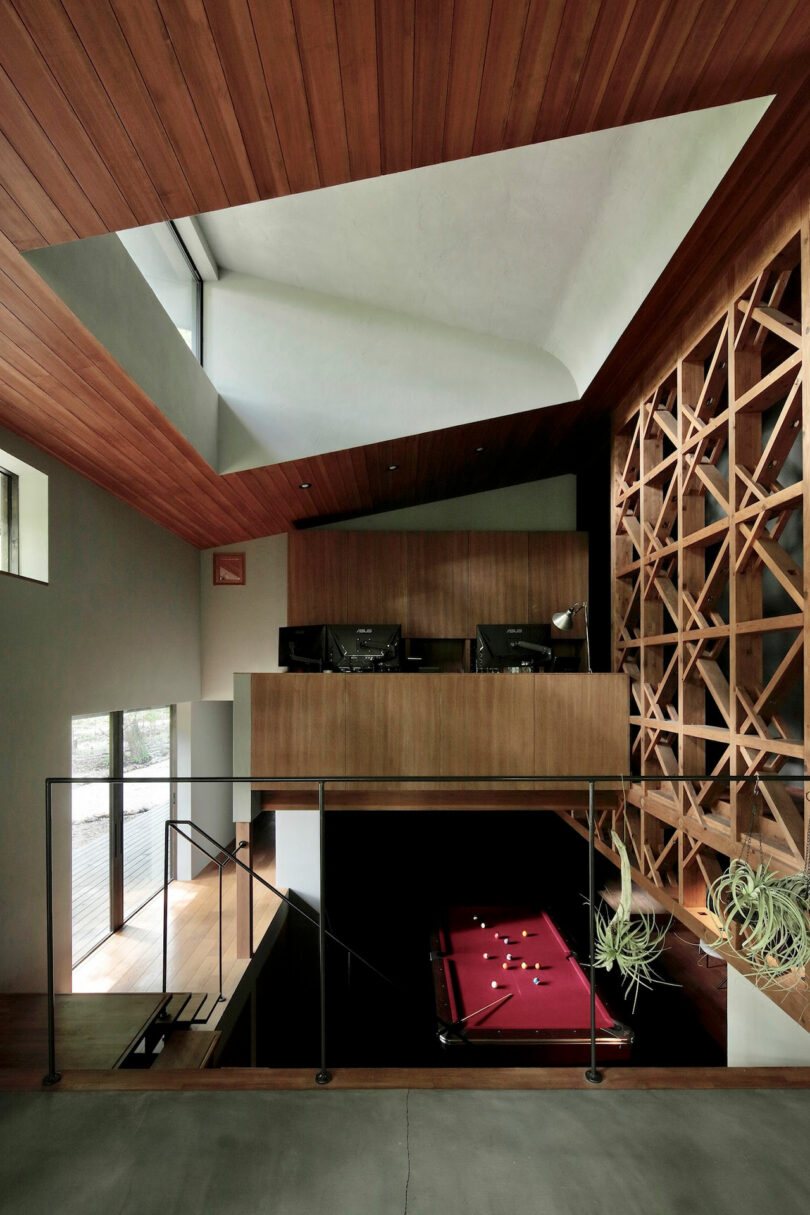 Modern interior with a wooden ceiling, mezzanine level, geometric wood wall, and a red pool table on a lower level, with natural light entering from large windows.