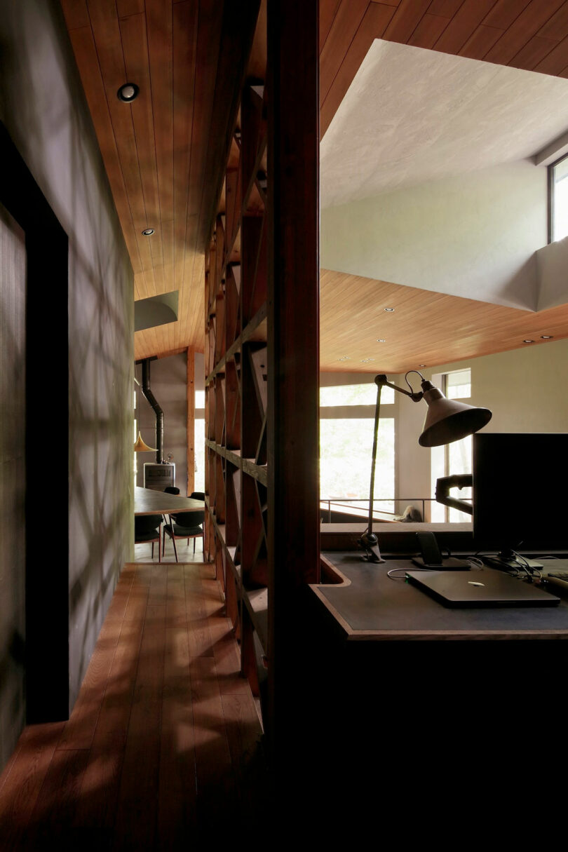 A modern interior with wood flooring and ceiling, a built-in bookshelf, a hallway, a desk with a lamp, and large windows letting in natural light.