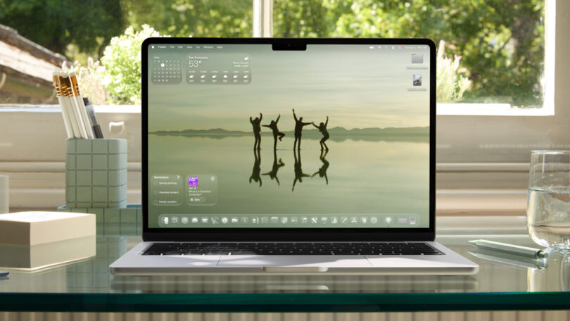 A laptop on a glass desk displays a desktop with a scenic wallpaper of people silhouetted against water, surrounded by desktop widgets and icons.