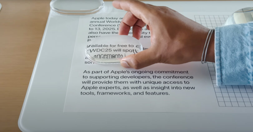 A hand holds a magnifying glass over printed text, enlarging part of a paragraph about Apple’s annual Worldwide Developers Conference.