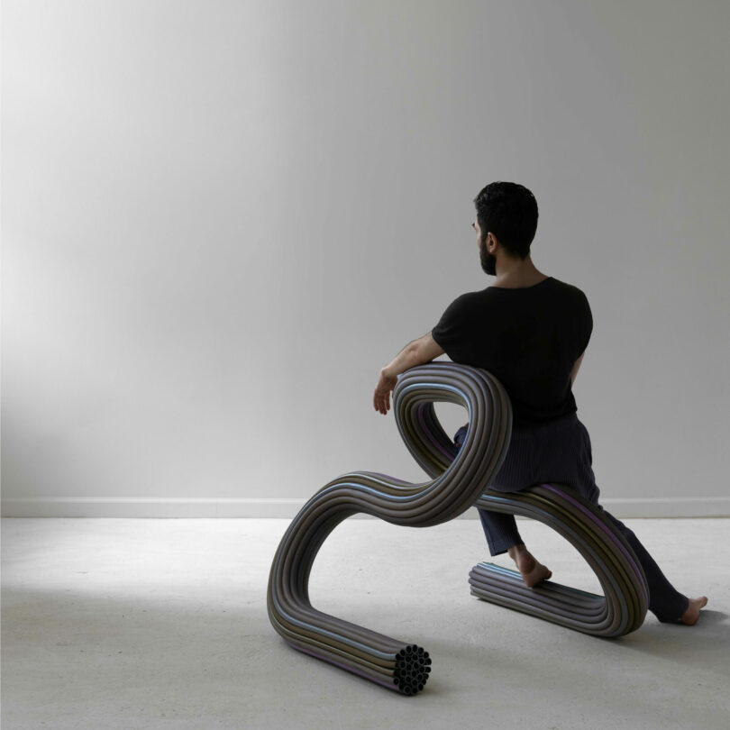 A person sits on a modern, sculptural ladder made of curved, multicolored tubes in a minimal, empty room with a white wall