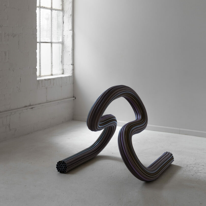 A sculptural artwork made of multiple curved, tubular rods is displayed on a white floor in a minimal, naturally lit room with a large window.