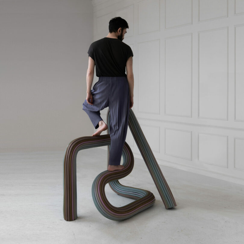 A person stands barefoot on an abstract, multicolored, looped stepladder in a minimal room with white paneled walls