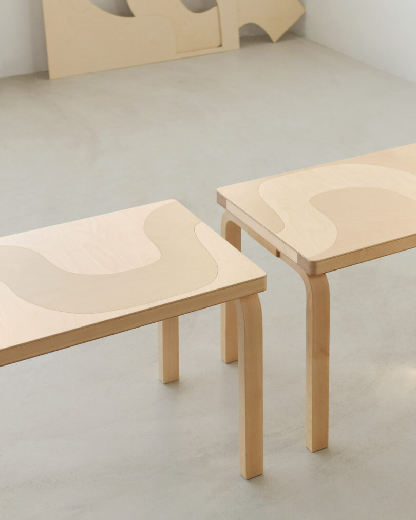 Two light wood tables with curved inlaid patterns on their surfaces are placed on a smooth, light-colored floor in a minimalist room