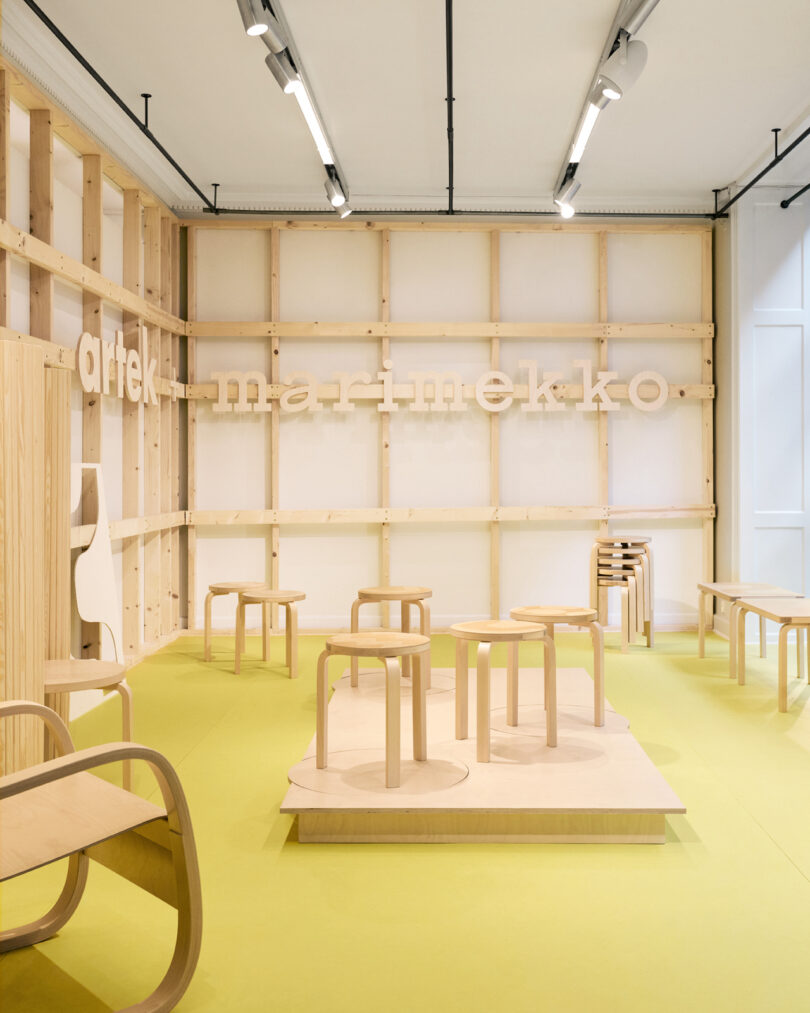 Minimalist showroom with light wood stools and chairs on a yellow floor, featuring “artek marimekko” signage on an unfinished wooden wall