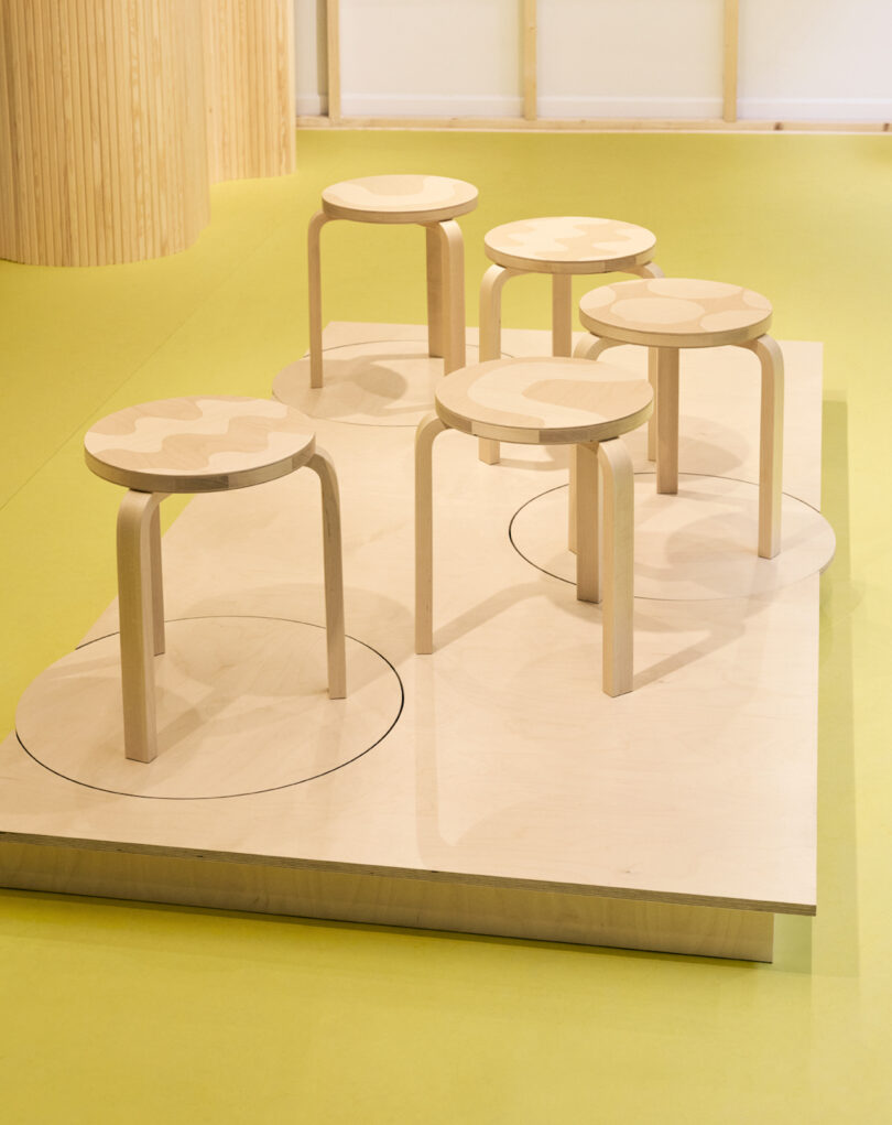 Five light wooden stools with patterned seats are displayed on a raised wooden platform with circular outlines, set against a yellow floor