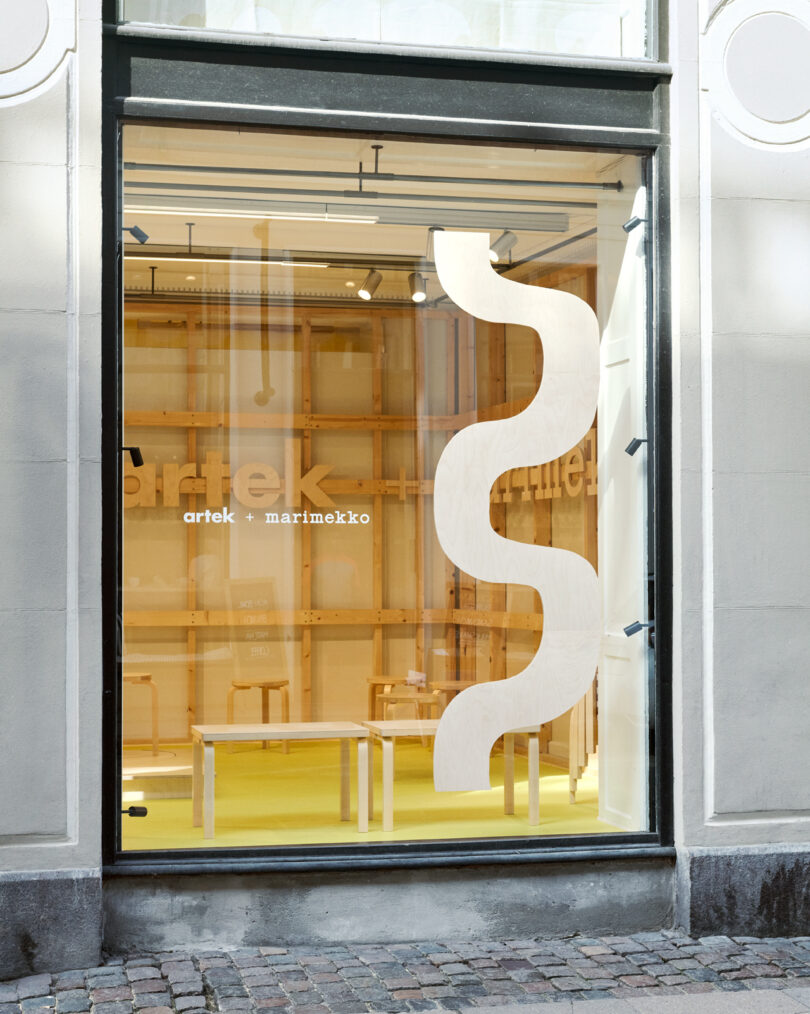 Storefront window featuring a large, wavy wooden panel and interior with wooden furniture; "artek" and "marimekko" logos visible on the glass