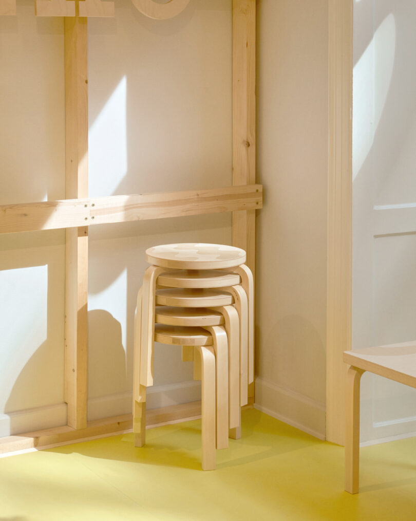 Four light wood stools are stacked neatly in the corner of a room with light-colored walls and yellow flooring