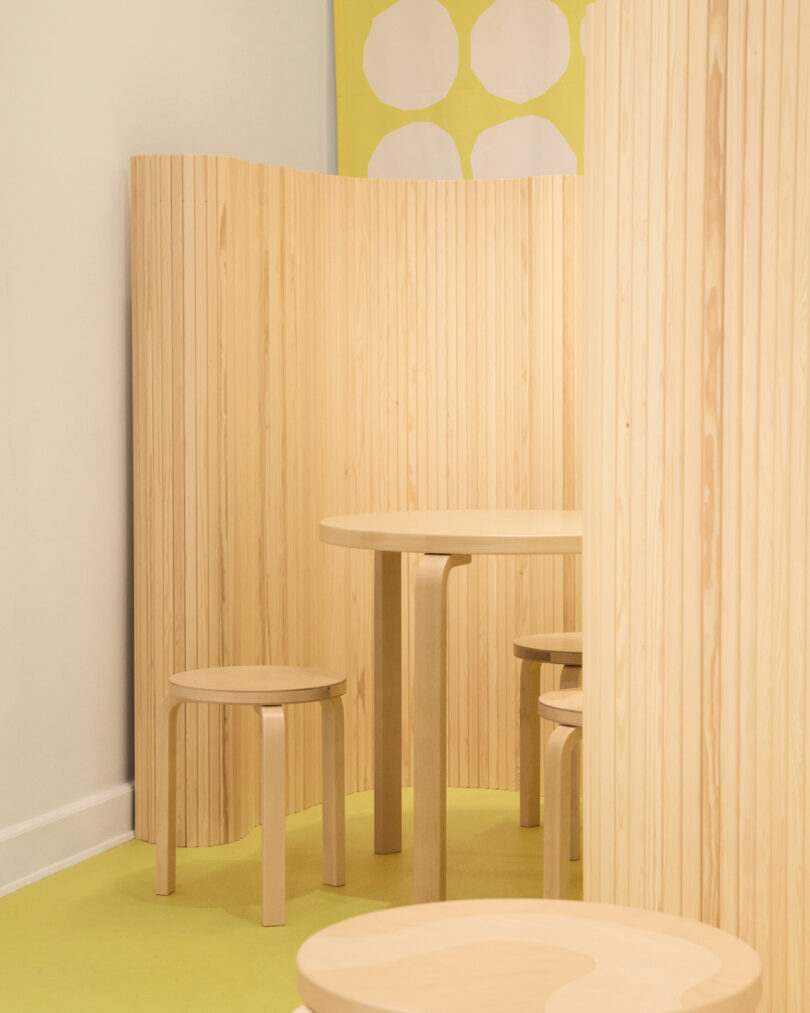 A minimalist room features light wood furniture, including a round table and stools, with vertical wood paneling and a yellow floor