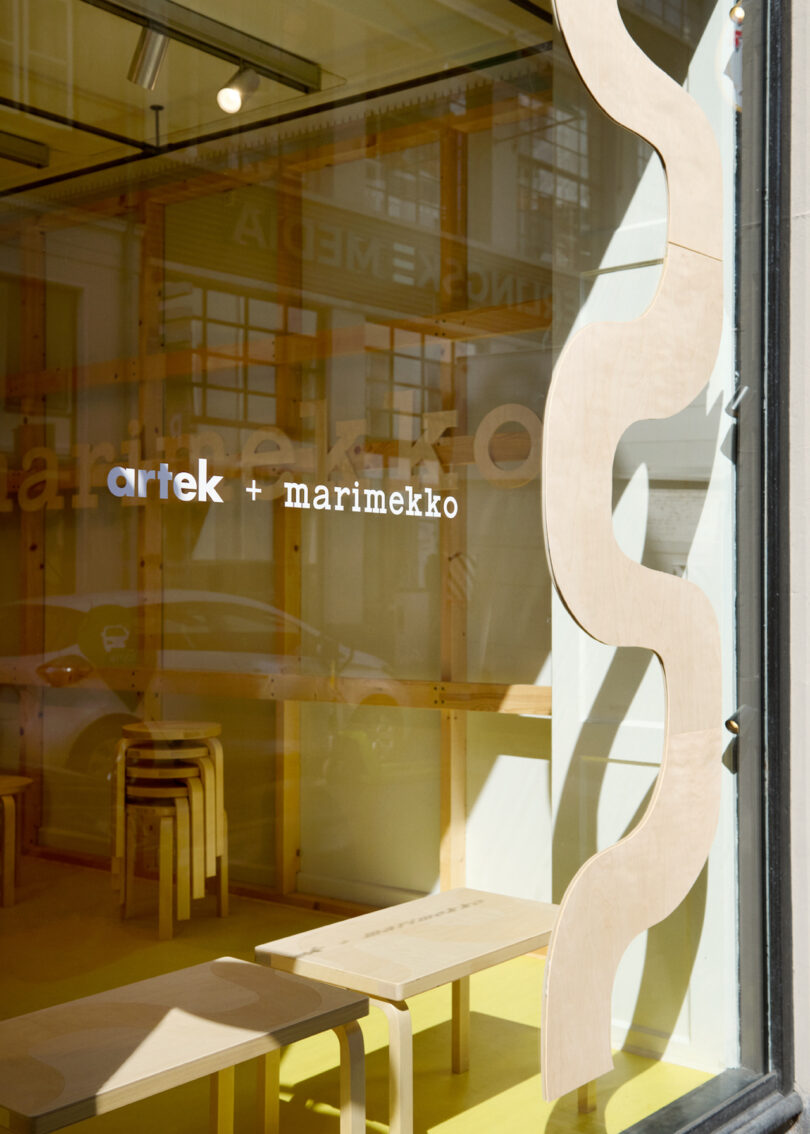 Interior of a storefront with wooden stools, a wavy wooden panel, and "artek + marimekko" written on the window. Natural light fills the space