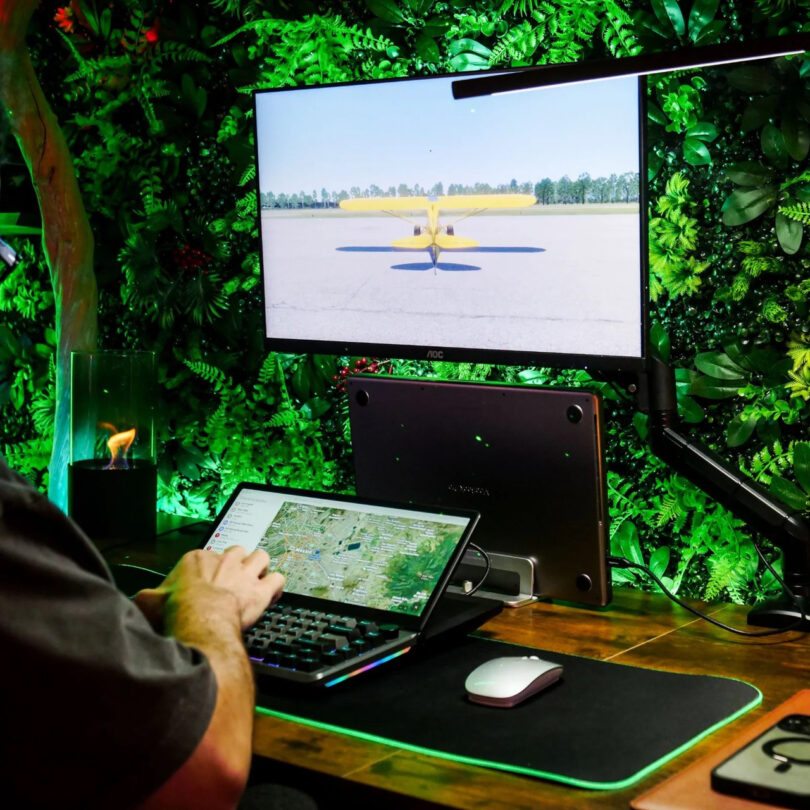 A person uses a keyboard and laptop at a desk with a glowing green backlight; a monitor displays a yellow airplane on a runway.