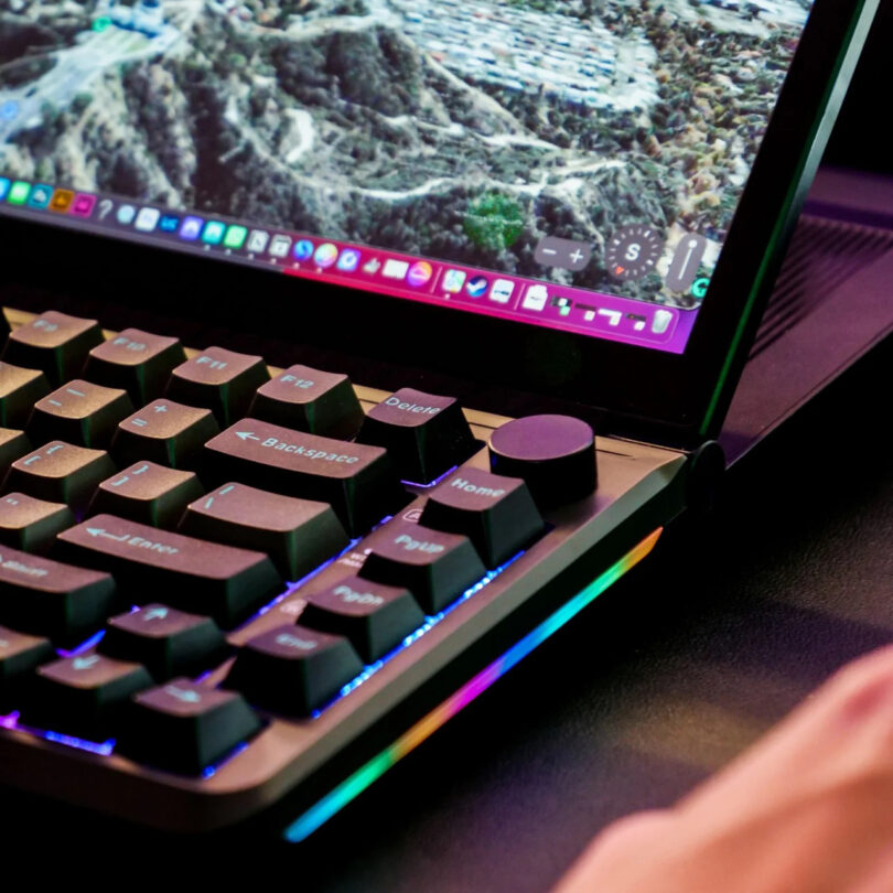 Close-up of a laptop keyboard with RGB backlighting, showing part of the screen displaying a landscape image and application icons on the taskbar.