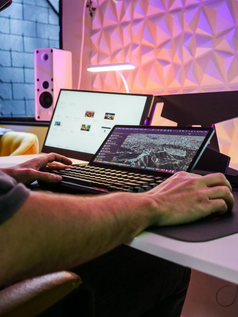 Person using a keyboard and mouse at a desk with two monitors; one displays photos, the other shows a map. Modern workspace with colorful lighting and geometric wall panel.