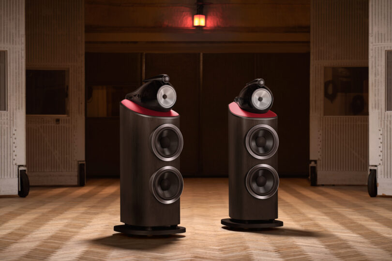 Two high-end Bowers & Wilkins floor-standing speakers with three front-facing drivers and a red top are positioned on a patterned wooden floor in a dimly lit room.