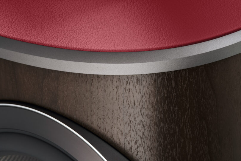 Close-up of a Bowers & Wilkins object with a red leather surface, a brushed metal edge, and wood grain finish below, featuring part of a circular design detail.
