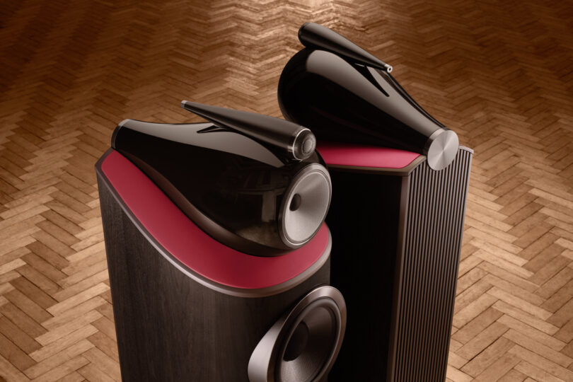 Two high-end Bowers & Wilkins black speakers with red accents are positioned on a herringbone wood floor, photographed from above.