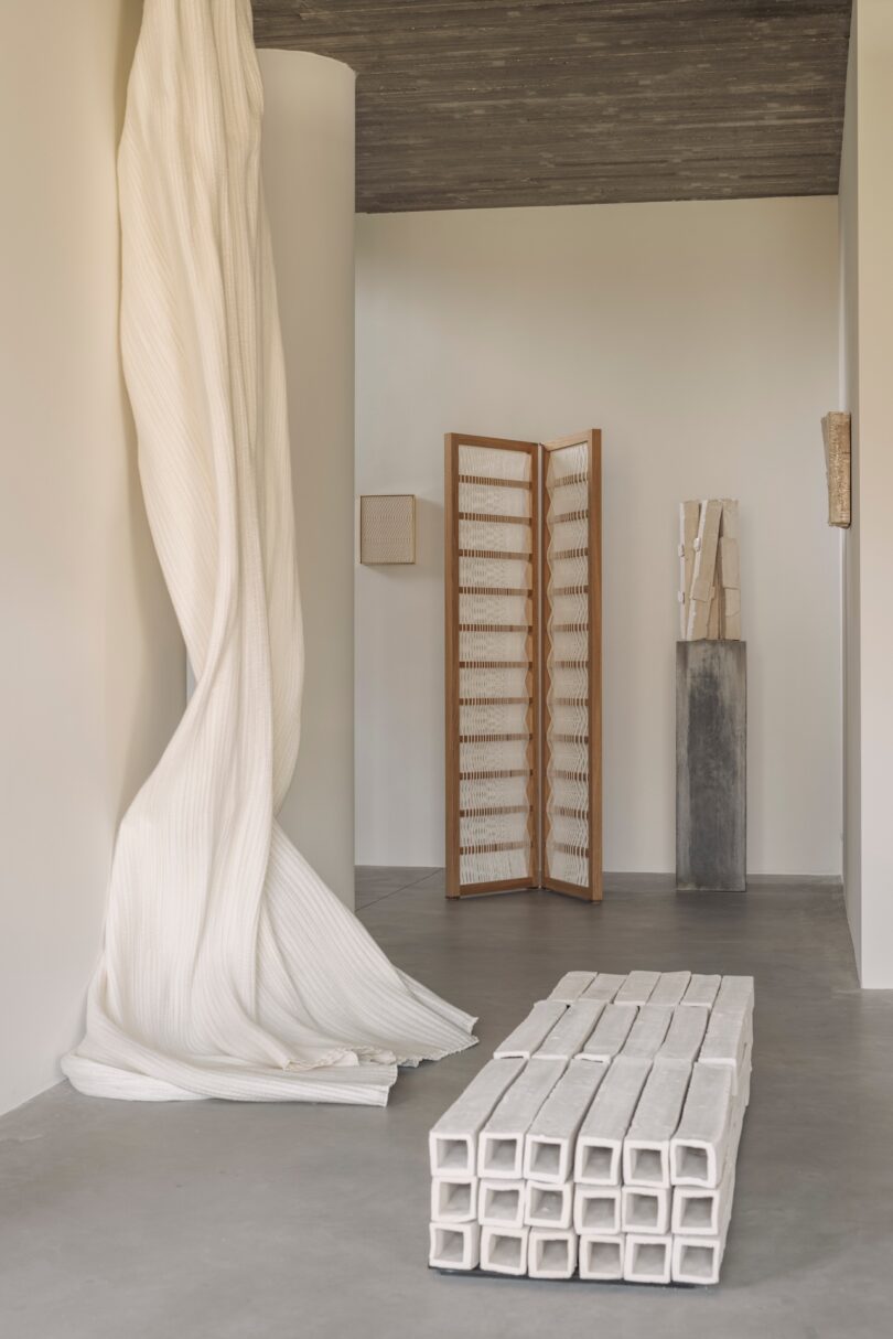 A minimalist room features a tall draped white fabric, a wooden folding screen, a grid-like bench, and textured sculptures on a polished concrete floor.