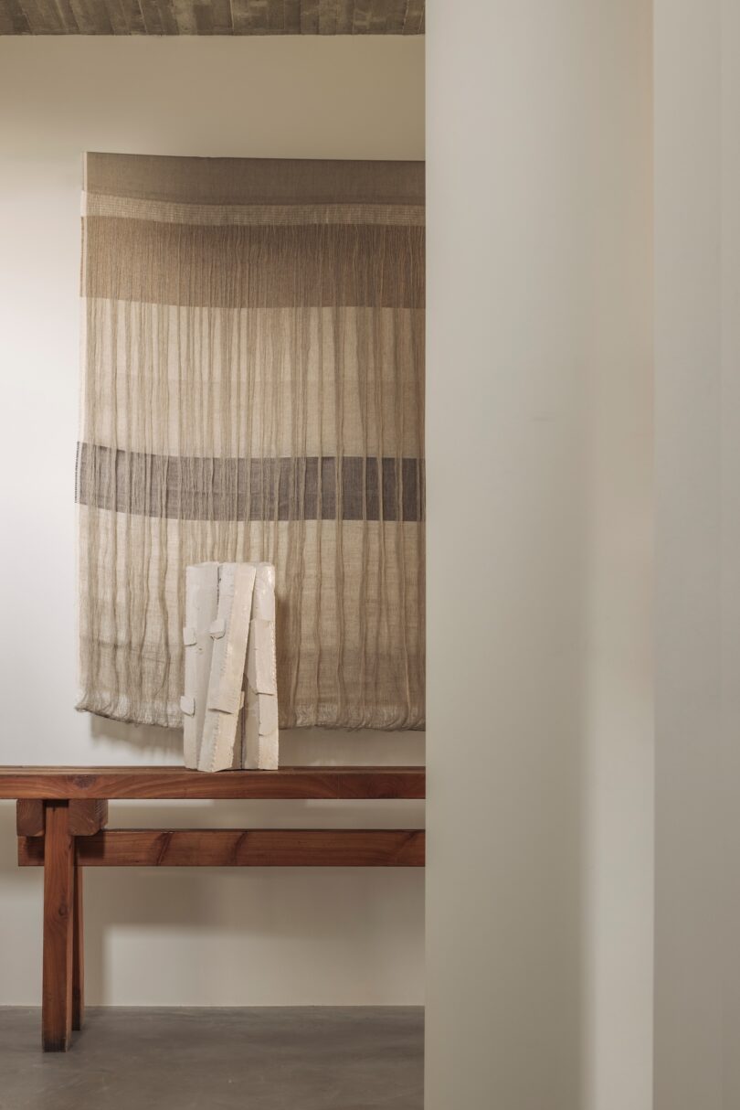 A minimalist interior with a wooden bench, a textured wall hanging in neutral tones, and a white sculptural object in front of it.