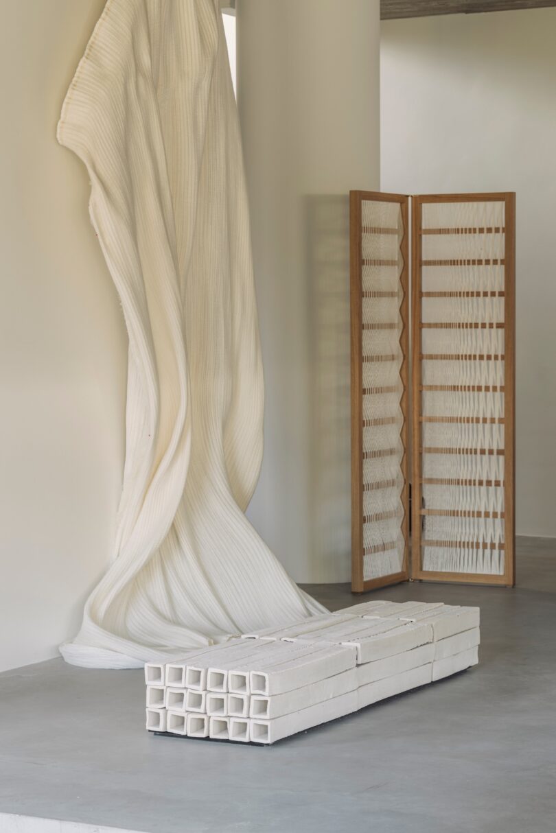 Minimalist room with a sculptural white fabric installation on the wall, a wooden folding screen with white panels, and stacked rectangular white blocks on the concrete floor.