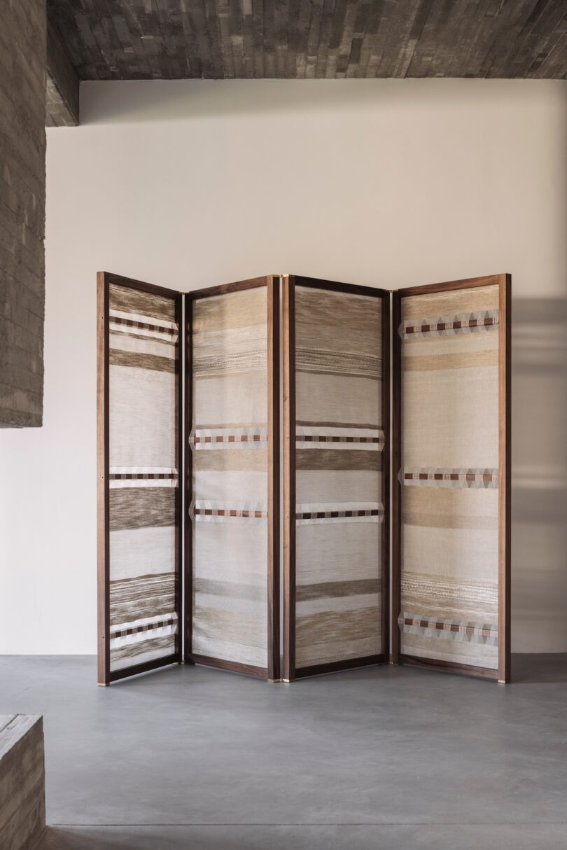 A four-panel wooden folding room divider with light-colored woven fabric panels stands in a minimalist room with concrete floors and ceiling.