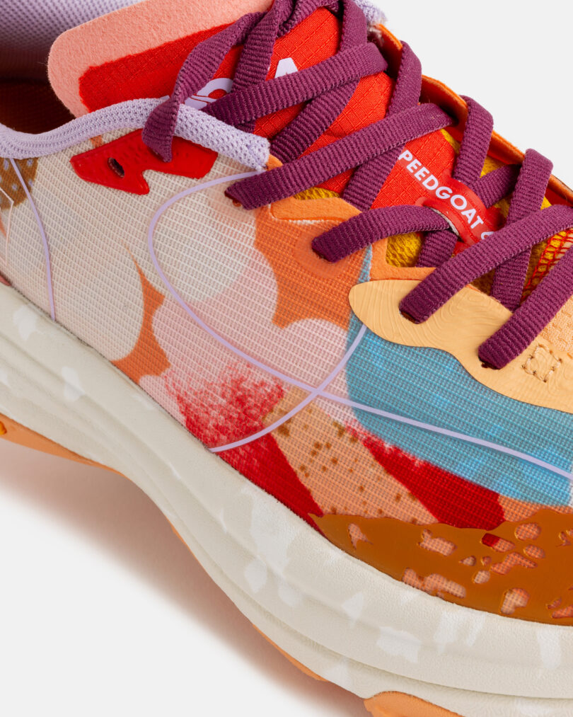 Close-up of a colorful, patterned running shoe with purple laces, orange and red overlays, and white midsole, showing the word "SPEEDGOAT" on the tongue