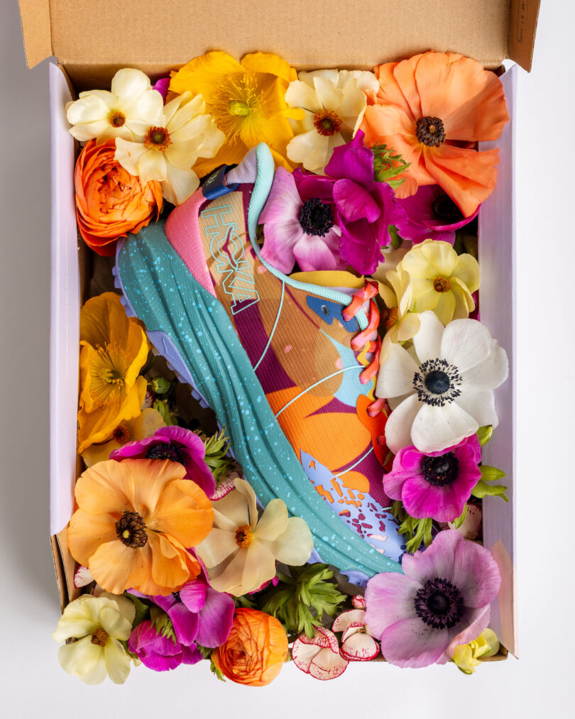 A colorful running shoe in a box surrounded by assorted vibrant flowers, including orange, yellow, and purple blooms