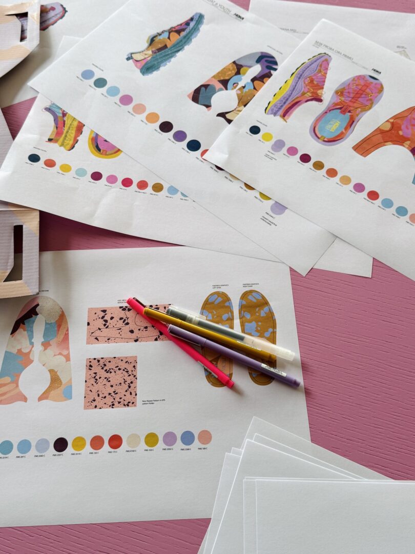 Several printed sheets with colorful sneaker design patterns, color swatches, and two pens lay on a pink textured surface