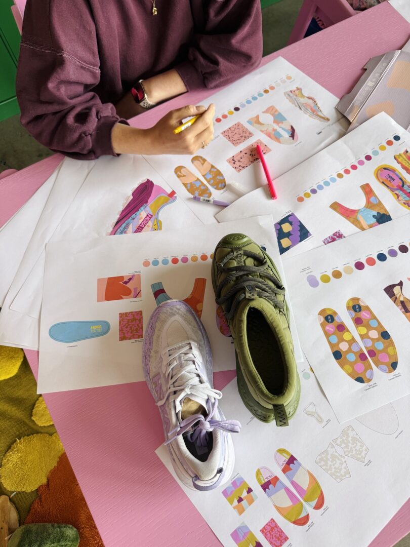 A person sits at a pink desk with sneaker design sketches, color swatches, a pen, and two sneakers—one white with purple accents, one green—placed on the papers