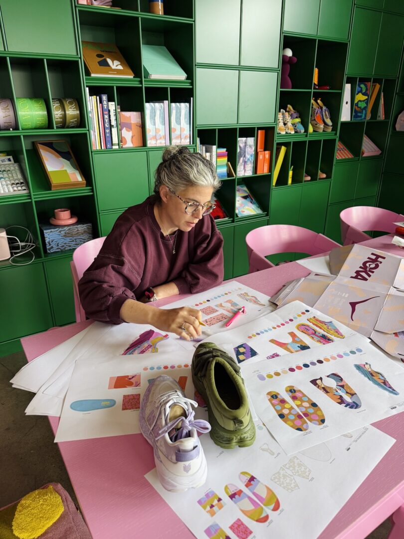 A person reviews colorful sneaker design sketches at a pink table, with two sample shoes and design materials, in a room with green shelving