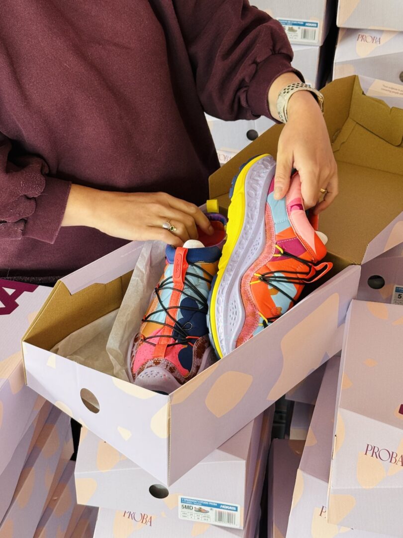 A person opens a shoebox containing a pair of colorful sneakers, surrounded by similar boxes